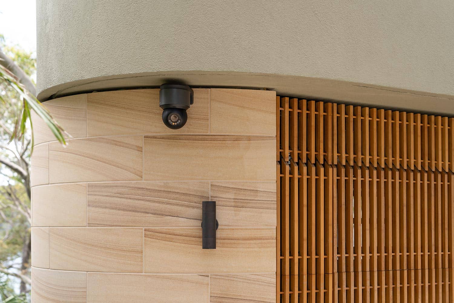 Black security camera and outdoor light fixture mounted on a curved tan stone wall