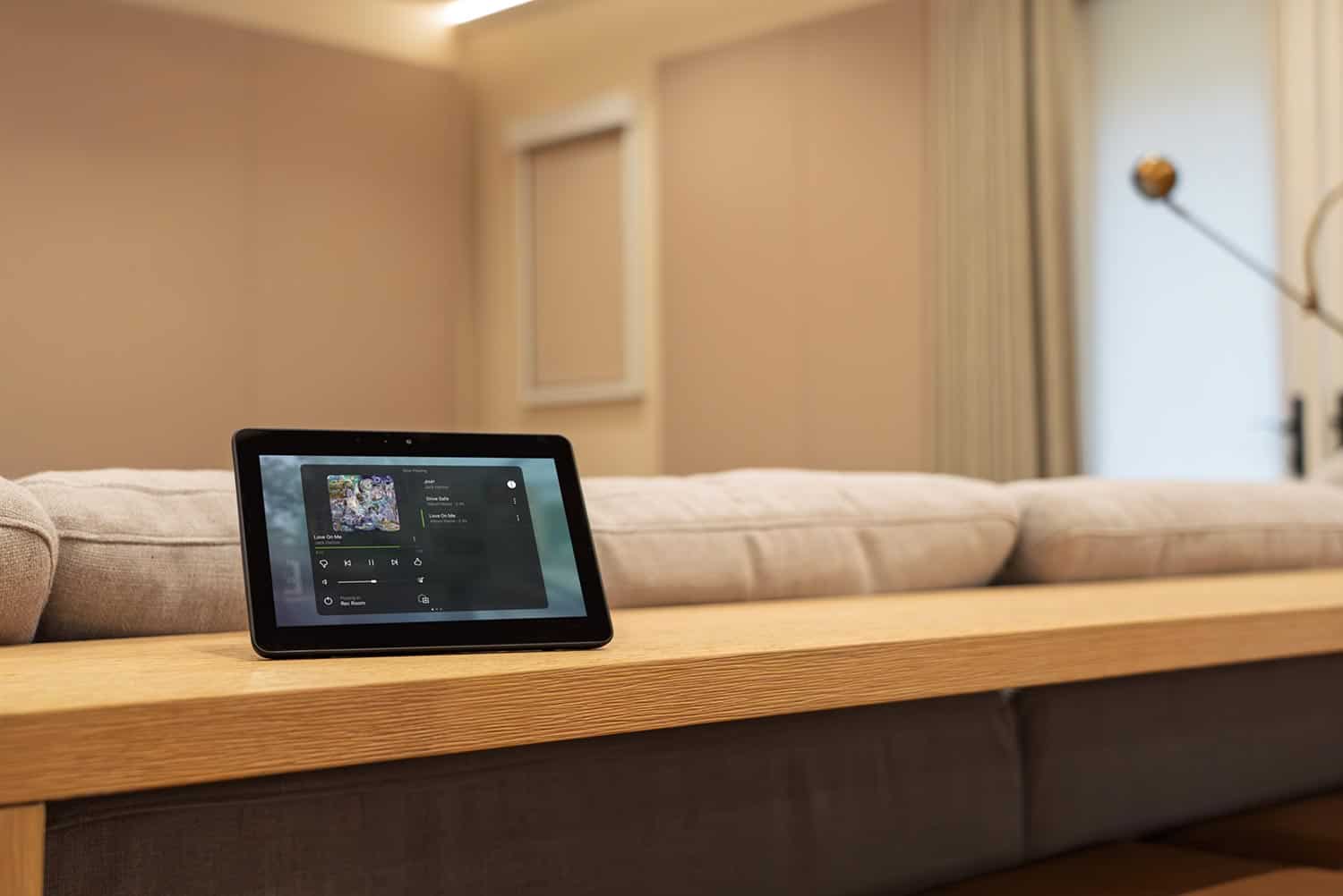 Portable smart home touch screen showing a music player on a light wood surface